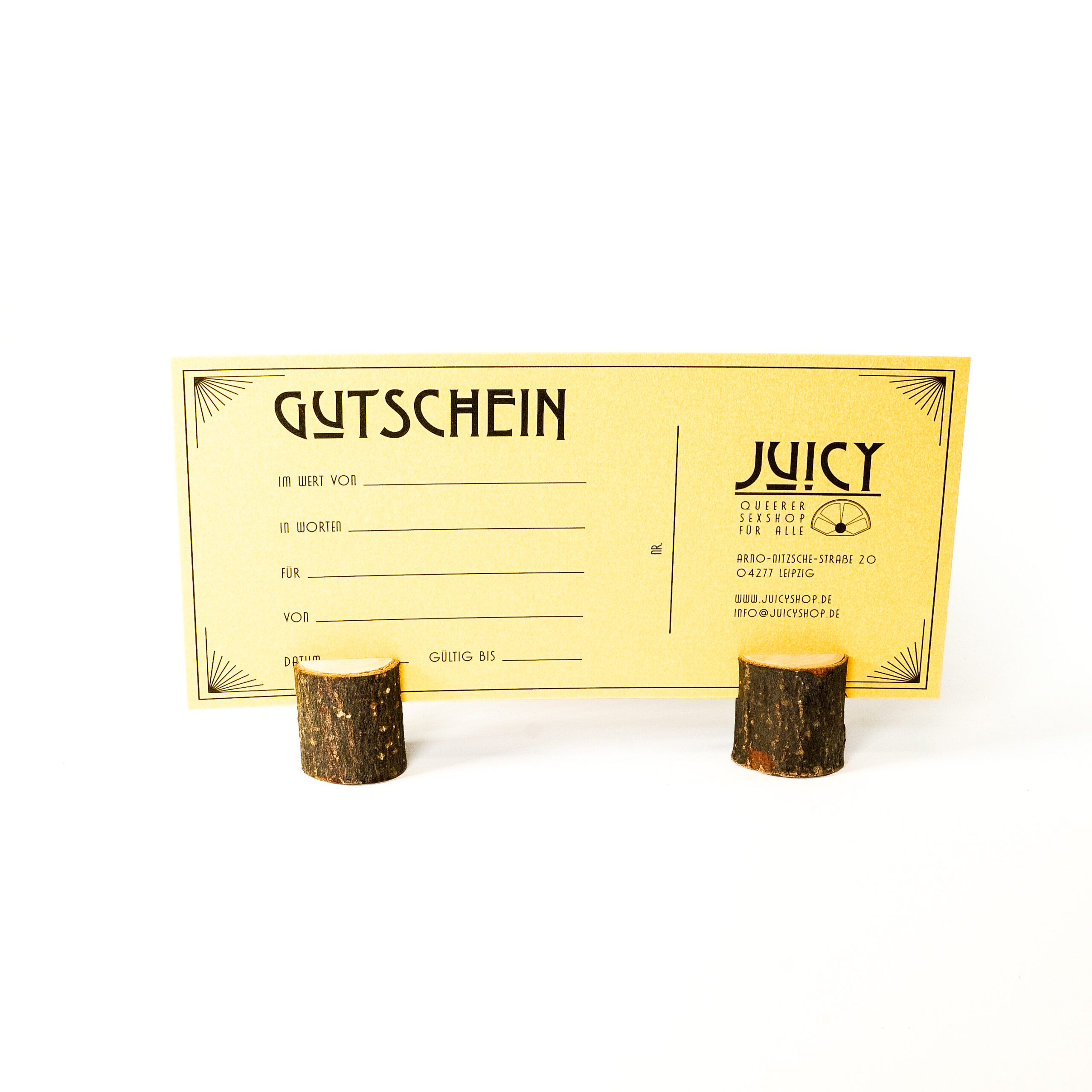 Produktbild: Ein goldener Gutschein steht auf kleinen Holzklötzen. Auf dem Gutschein steht die Adresse von Juicy, Zeilen für den Gutscheinbetrag und Zeilen von und für wen der Gutschein ist.