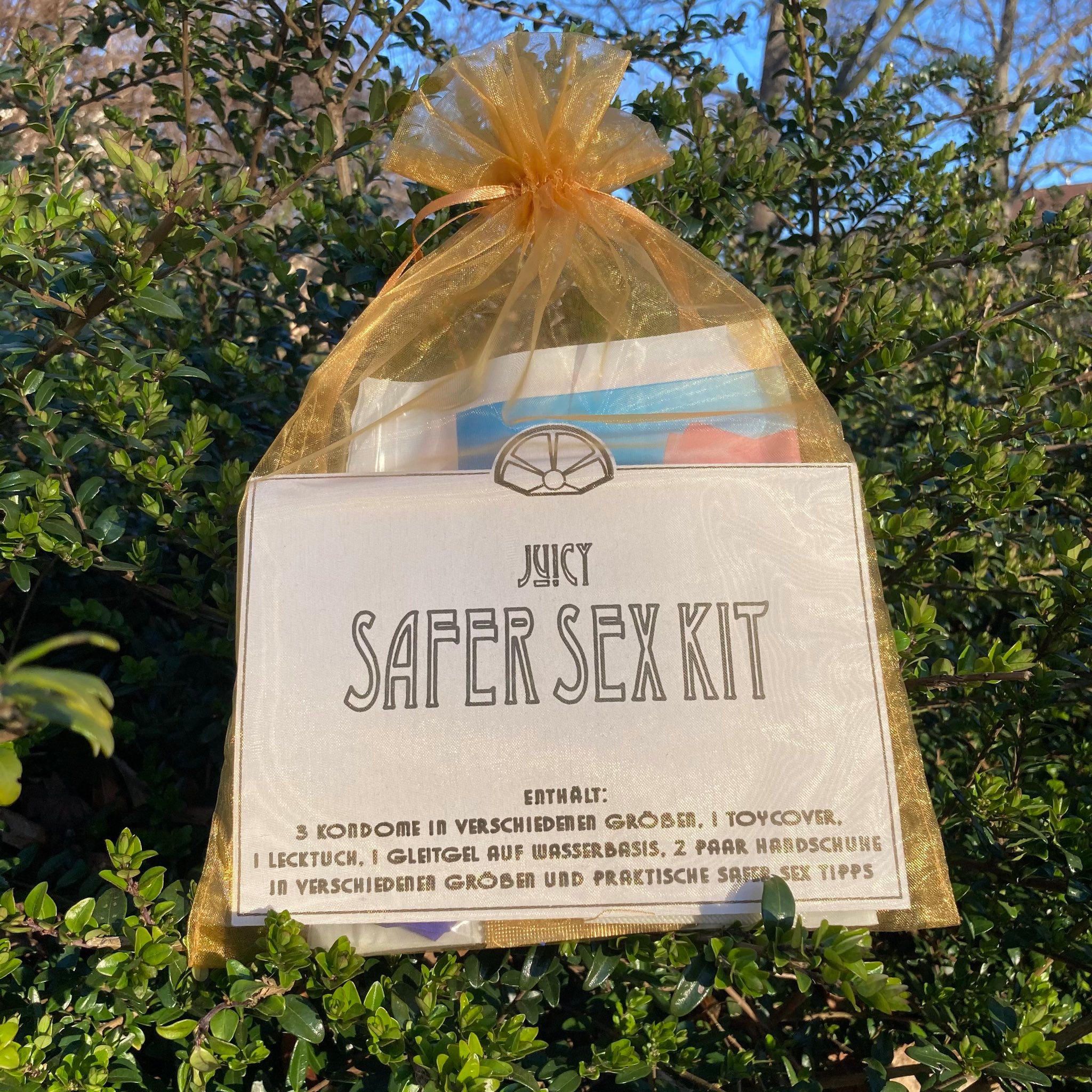Ein gold-transparenter Organza Beutel liegt in einem Busch. Darin sind verschiedene Safer Sex Utensilien zu sehen. Es liegt ein Zettel bei auf dem Safer Sex Kit steht.